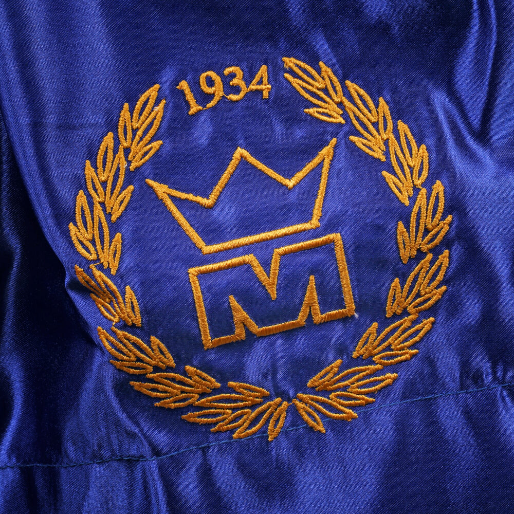Détail macro du blason doré à la couronne daté 1934, brodé sur la jambe du short de boxe Montana MUAY THAI NewCode en satin bleu.