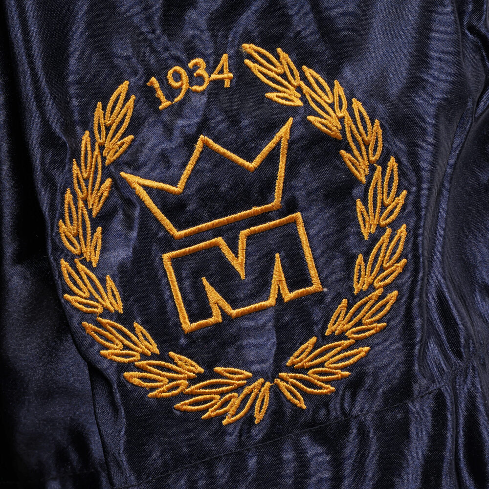 Détail macro du blason doré à la couronne daté 1934, brodé sur la jambe du short de boxe Montana MUAY THAI NewCode Poseidon en satin navy.
